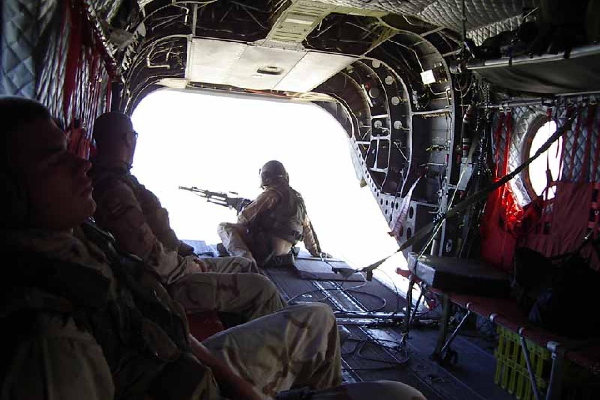 Drie soldaten in een Helicopter met de laadklep open. Soldaat met mitrailleur kijkt via laadklep naar buiten
